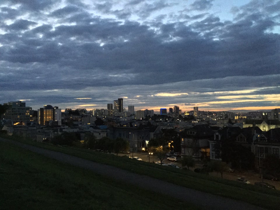 Bits of light blue sky peaking through mottled clouds that grew thicker towards the horizon, with just a few streaking cracks letting through yellow and orange light, the buildings in the distance mostly in darkness with their lights still on, streetlamps illuminating the street and houses below, a path barely visible, with green grass incline and decline on the sides.