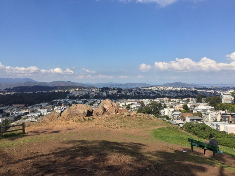 Northward view of Tank Hill Park with Marin hills, Golden Gate bridge, and Angel Island in the distance, largerocks and the bench at Tank Hill park in the foreground with one woman at each.