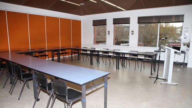 Room with orange wall, chairs and tables in a u shape, lecturn and microphone, and windows showing trees.