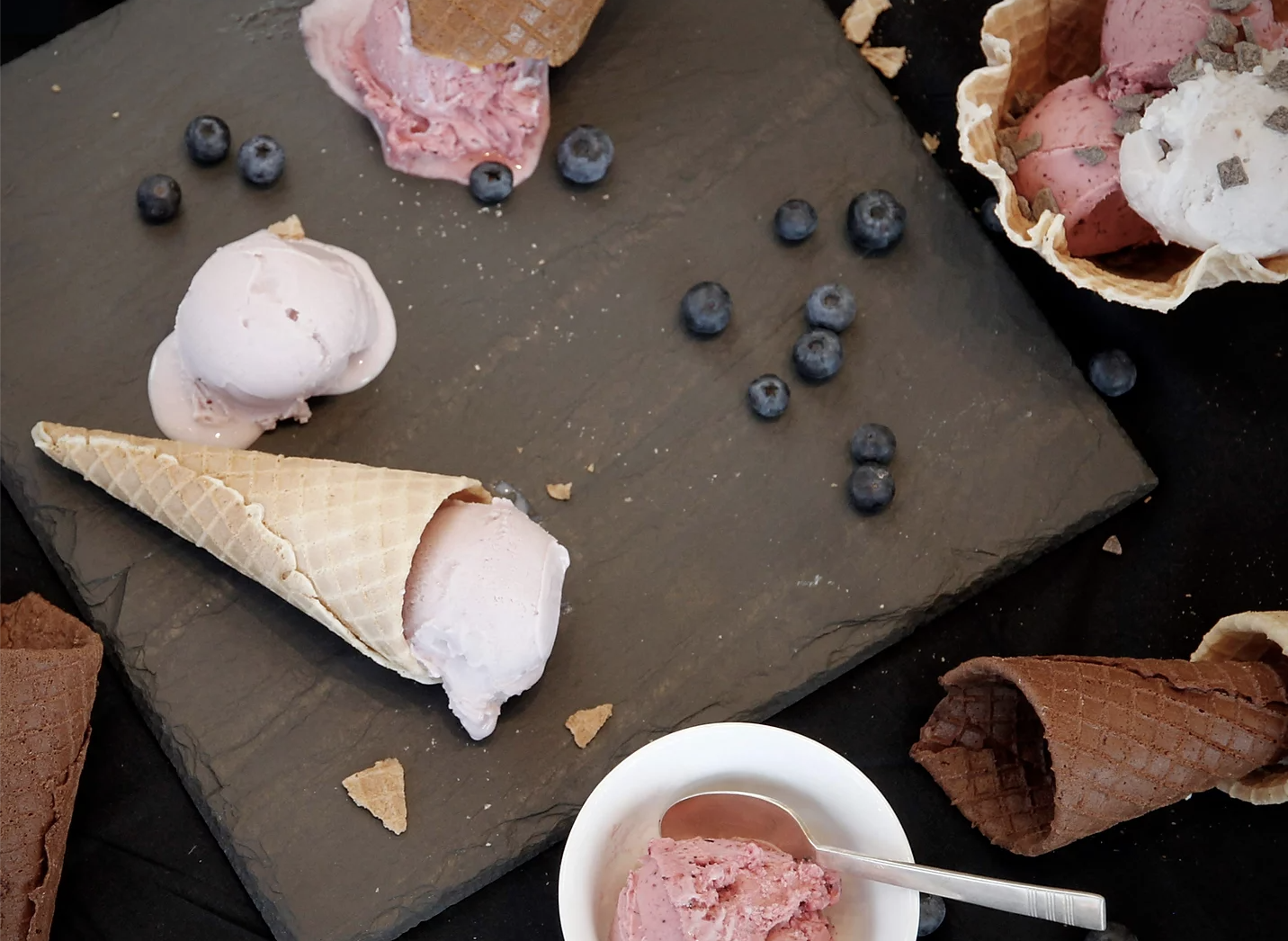 Gelato in cones and cups drips onto a wooden table.