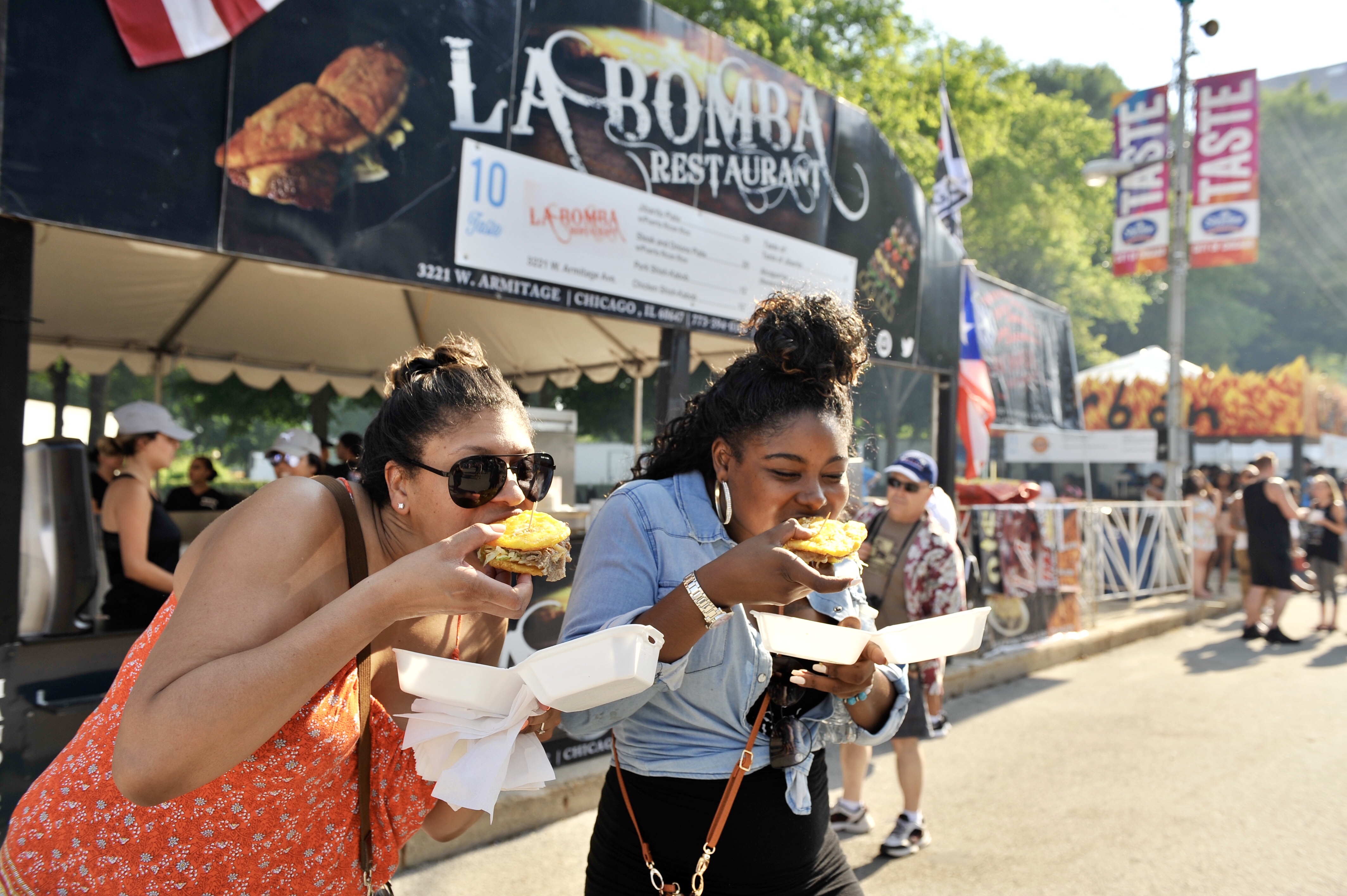 Two women bite into food standing outside.