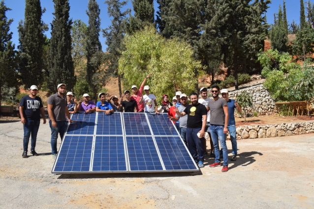 A group of people gathered around a small ground-mounted solar array, smiling at the camera.