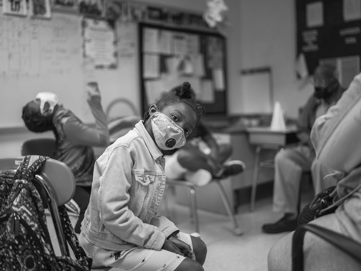 A young girl in a classroom sitting in a chair looking directly at the camera with her head tilted to the side