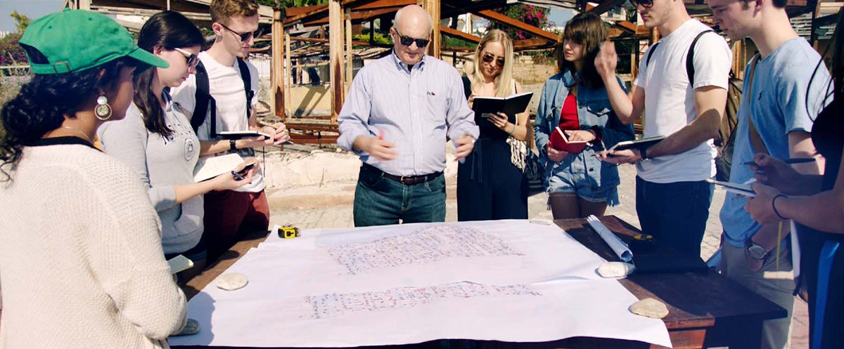 A man stands in the middle of a group of people surrounding a table with blueprints on it.