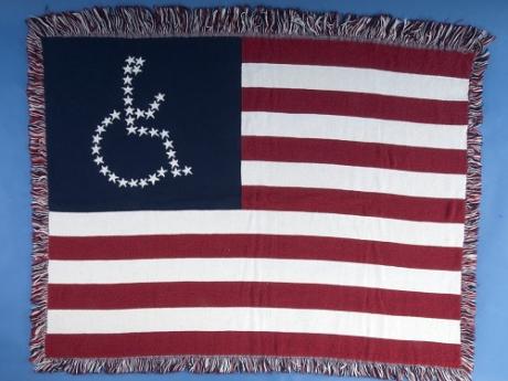 An image of an American flag woven lap blanket. Instead of the 50 white stars making up the upper left corner of the flag, the stars are arranged in the shape of the international symbol for accessibility- a figure in a wheelchair. 