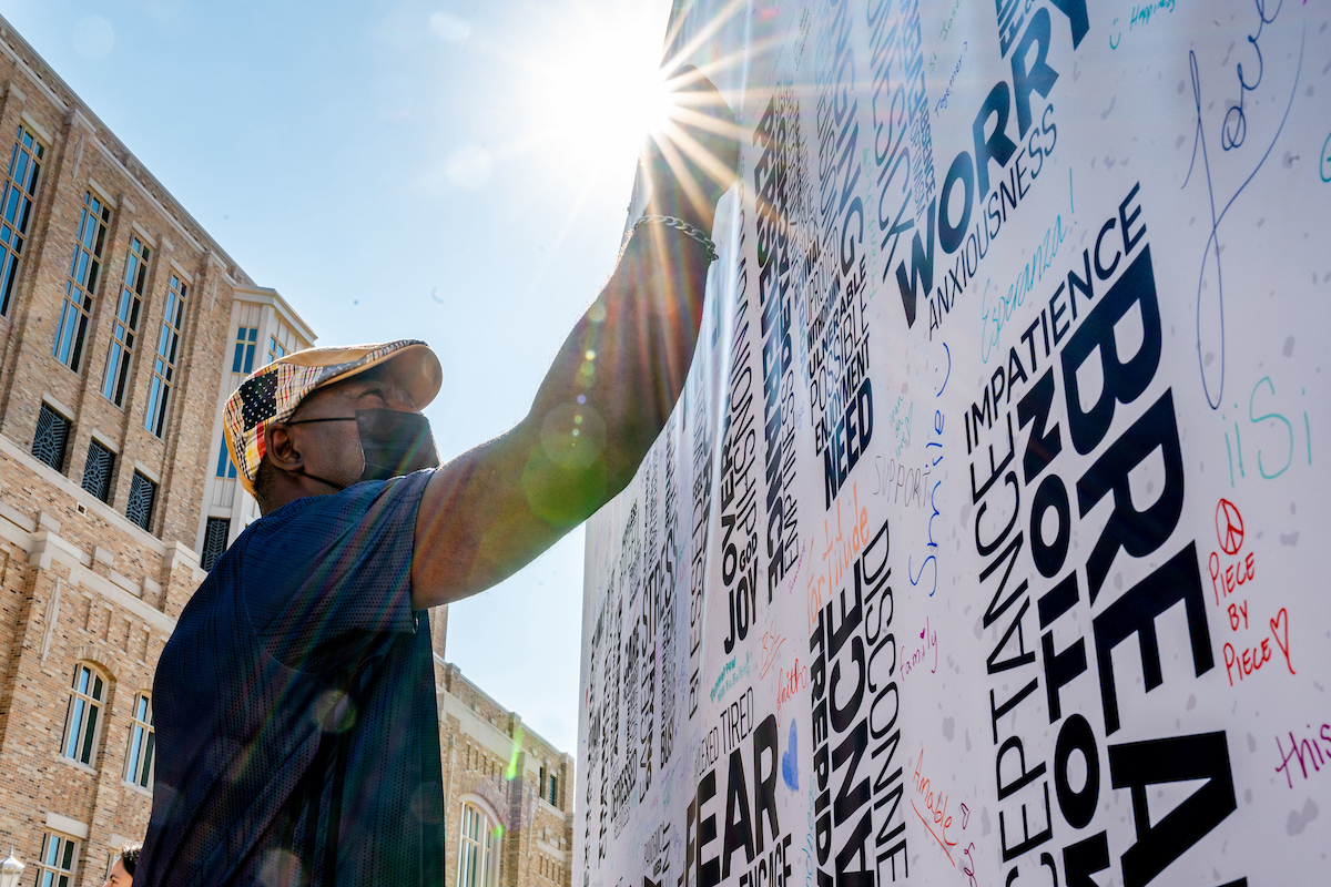 A man writes on a white billboard with a sun flare above him. The billboard has a word cloud of emotions students have felt this semester spelling out 'The Fighting Irish.'