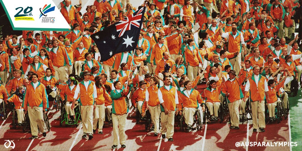 Australian Paralympic Team walking into the Opening Ceremony of the Sydney 2000 Paralympic Games being led by flag bearer Brendan Burkett waving the Australian flag. The team is dressed in beige pants and orange and green jackets. 