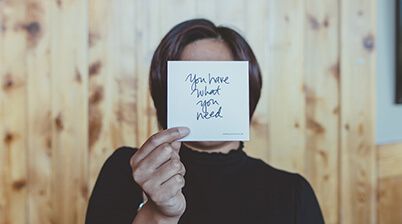 A person holding up a card that blocks their face. The card reads 'you have what you need'
