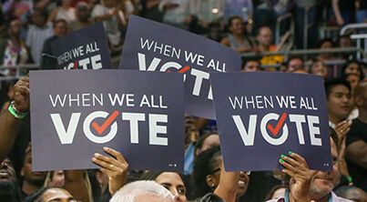 A crowd at a voter rally, several holding sign with the logo 'We All Vote