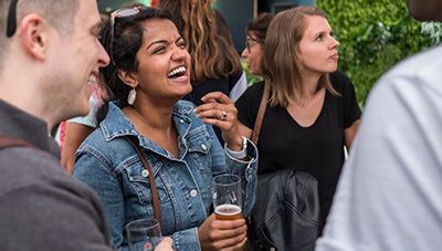 A woman with a beer laughing at a joke at an outdoor bar