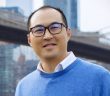 A man wearing black glasses and a blue sweater smiles in front of a bridge.