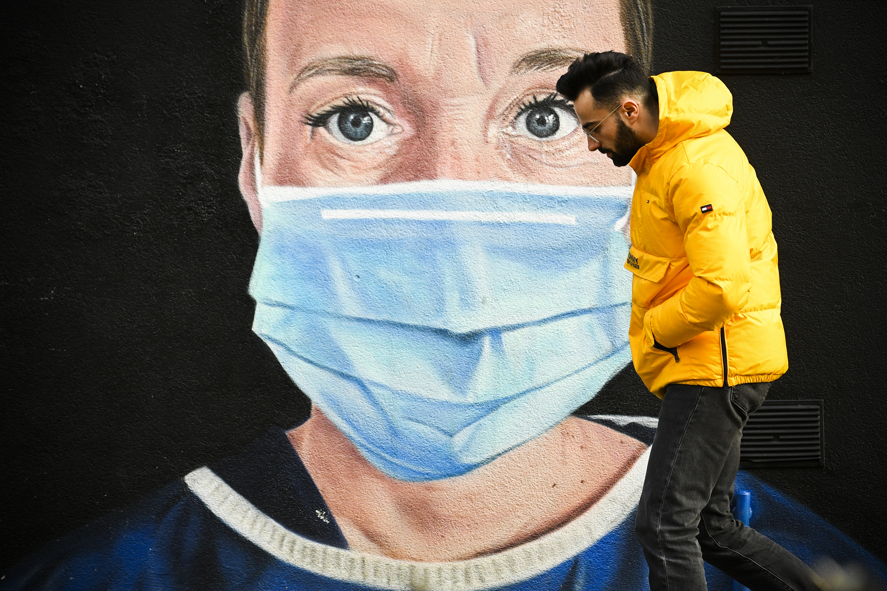 A person walks past a large mural of a face wearing a breathing mask.