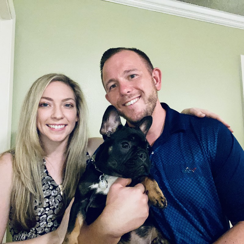 A man and a woman smile for a photo while holding a black French bulldog.