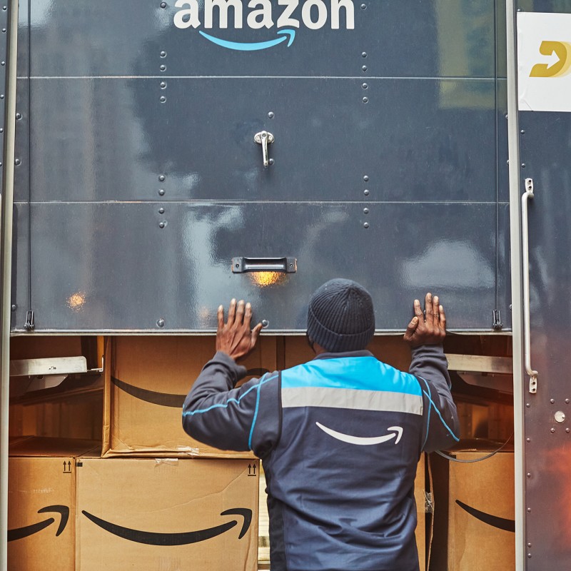 An image of a man closing the back of an Amazon delivery truck that is loaded with Amazon boxes inside of it.