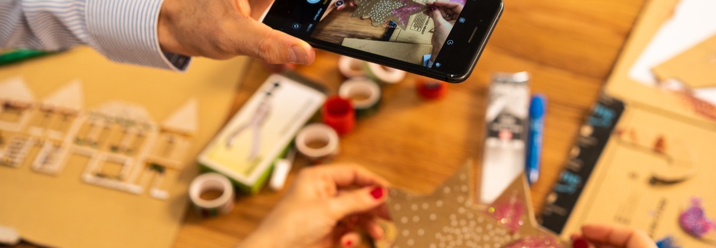 En una mesa de madera del salón, un primer plano de las dos manos de Marta con las uñas pintadas de rojo y la mano izquierda de Fernando sacando una foto con el teléfono. El va con una camisa de rayas blancas y azules. De fondo material para hacer manualidades. 