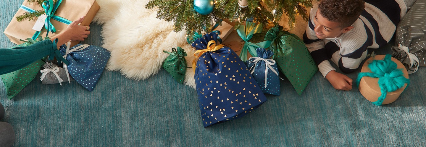 En los pies del árbol de Navidad se encuentra un niño de rodillas y lleno de bolsas de regalo de Amazon. En el suelo hay una alfombra de color verde-azul.  En el lado derecho está un niño moreno con un jersey de lana a rayas negras y blancas.  Al lado izquierdo se ve un brazo de una personas más mayor con un jersey verde. El árbol tiene un protector de color blanco y los paquetes de regalos son azules y verdes con motivos dorados.