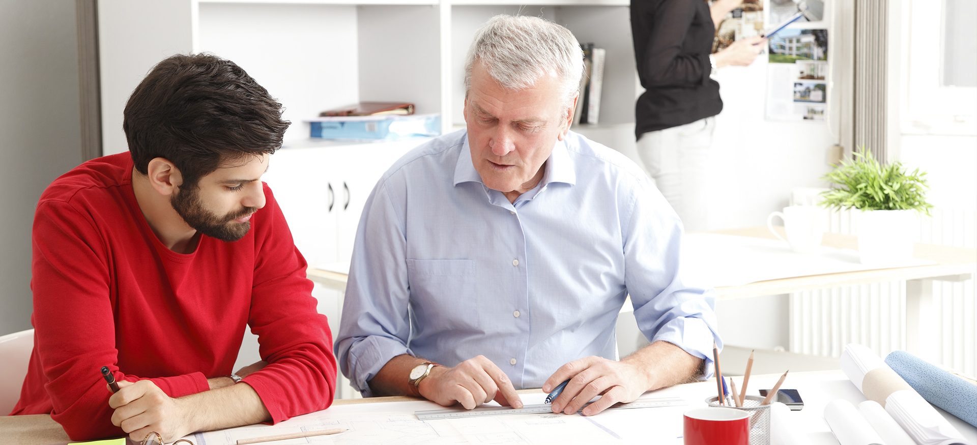 Two men are sitting at a white desk reading through documents on the table