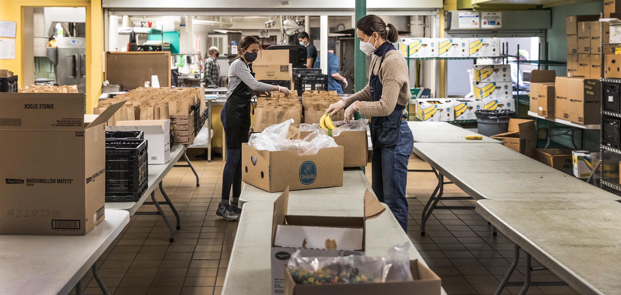 Multiple people unpack cardboard boxes and package brown-bag lunches