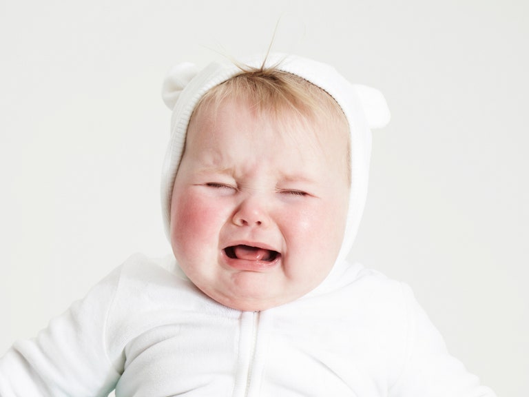 A crying baby in a white onesie in front of a white wall