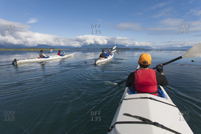 A small group of people kayaks in pristine waters of an inlet on the Alaska coastline.