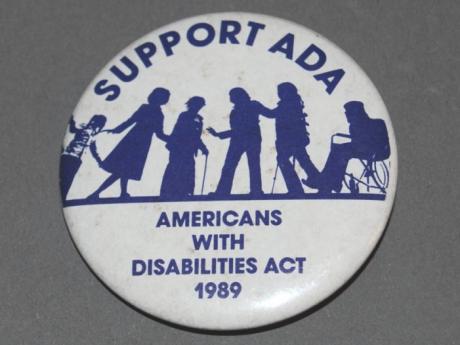 A white button with blue text reading: Support the ADA, Americans with Disabilities Act 1989. Between the two phrases in the middle of the button are silhouettes of six individuals some of whom are using mobility devices. 