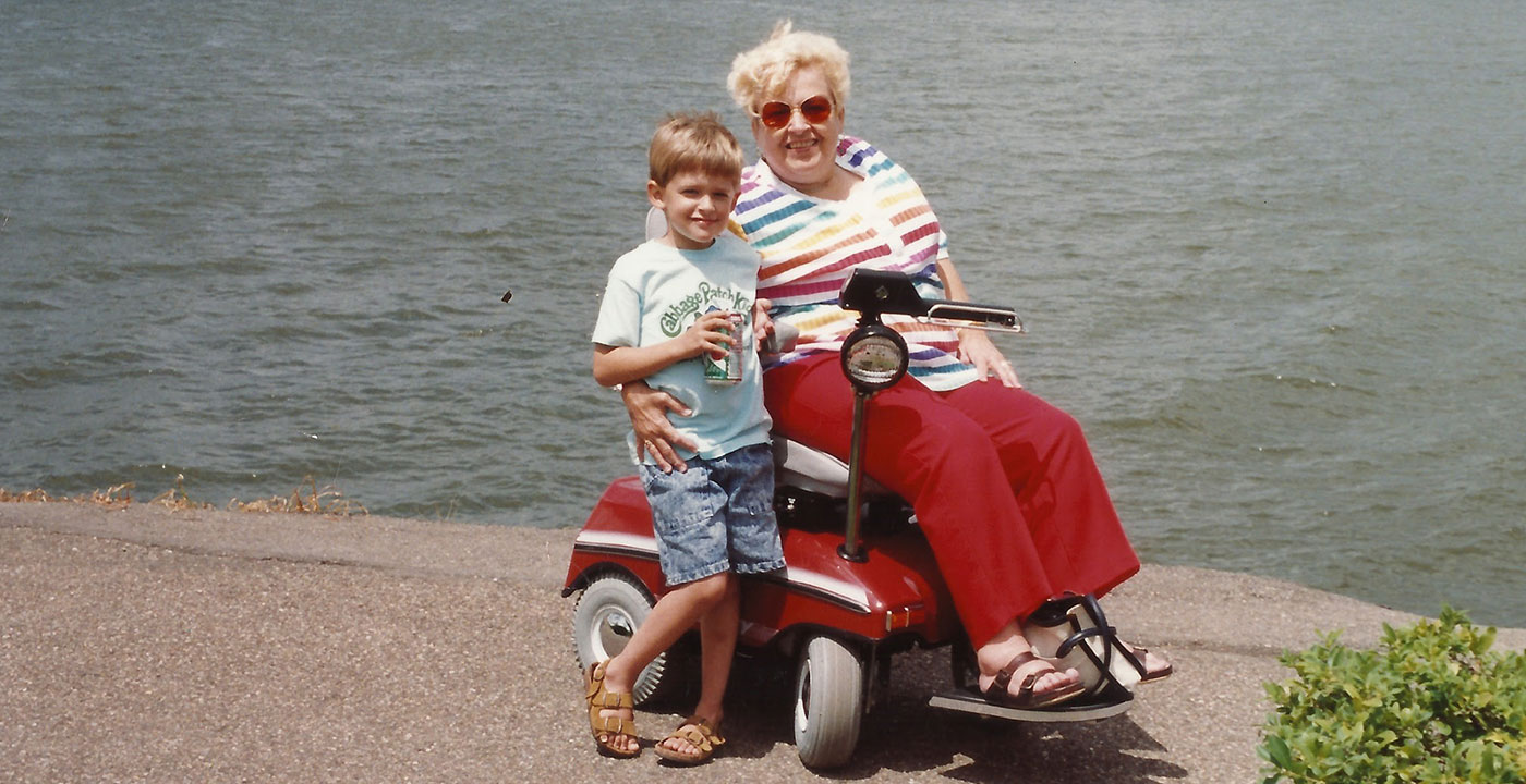 Old color photo of a young boy standing next to an older woman on an electric mobility scooter in front of a lake.