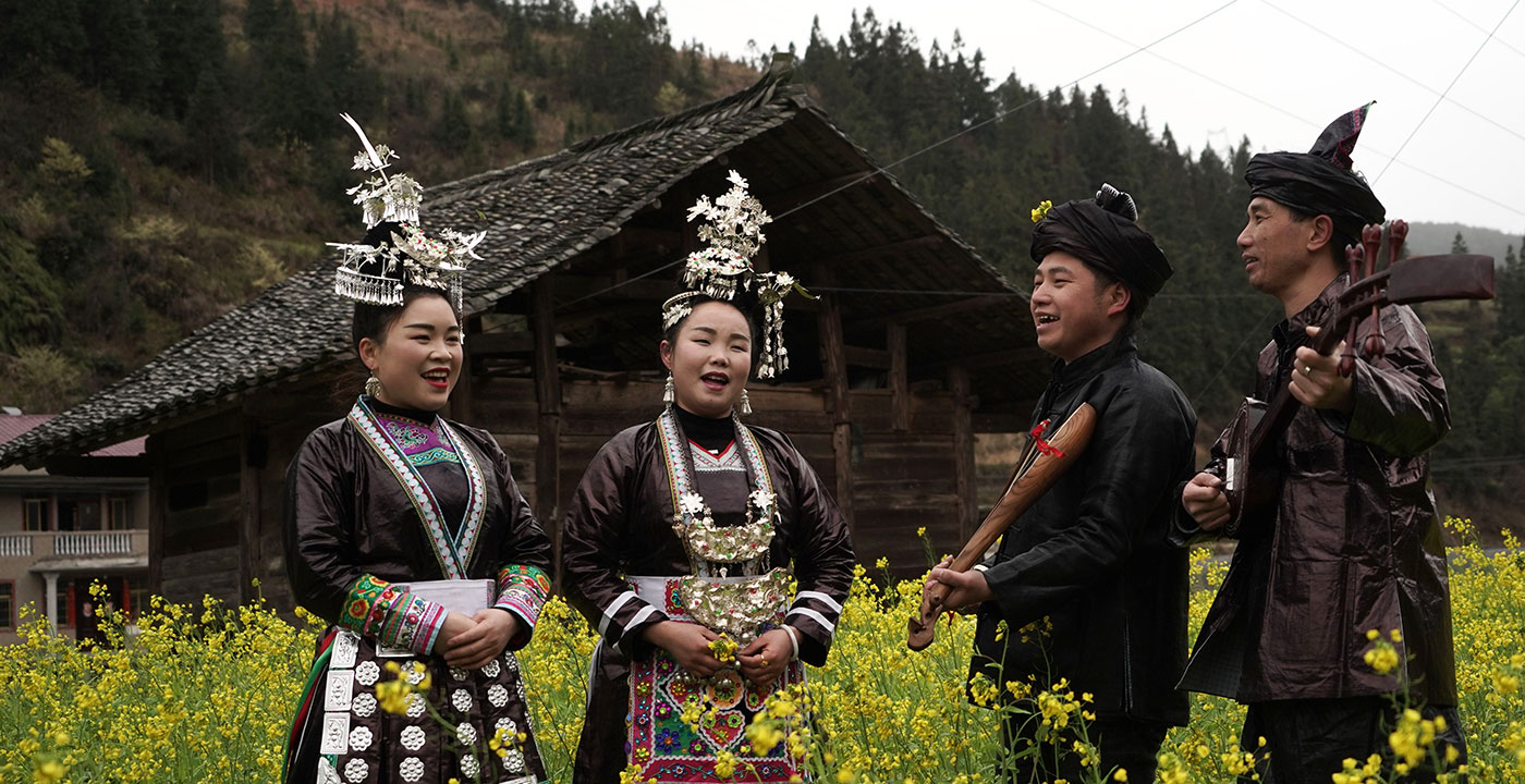 Two women singers and two male instrumentalists perform in ornate dress, outdoors in a field of tall yellow flowers.