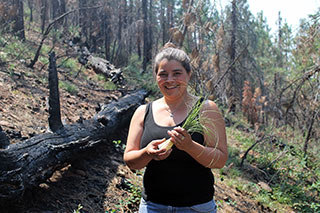 A young women holds a tuft of long grass, smiling on a forested hillside that looks like it's recovering from a fire.