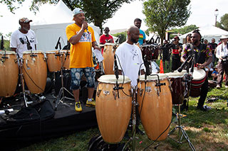 Man addresses an outdoor crowd on microphone as other men on congas around him prepare to perform.