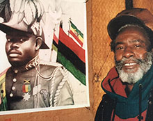 A Black man smiles, posing next to a poster of a Black man in military dress and the Pan-African flag.