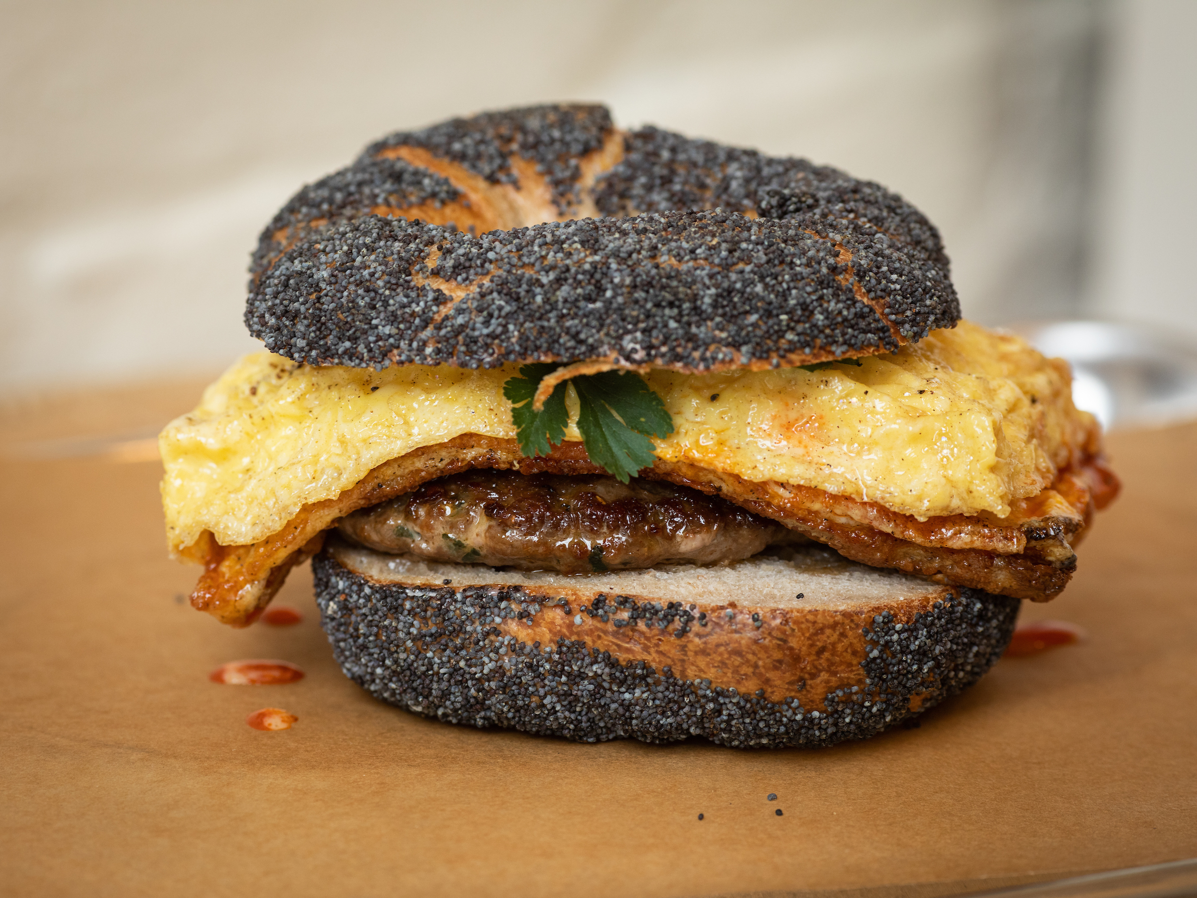 A bagel coated in sesame seeds is topped with egg, parsley, and a glistening sausage patty