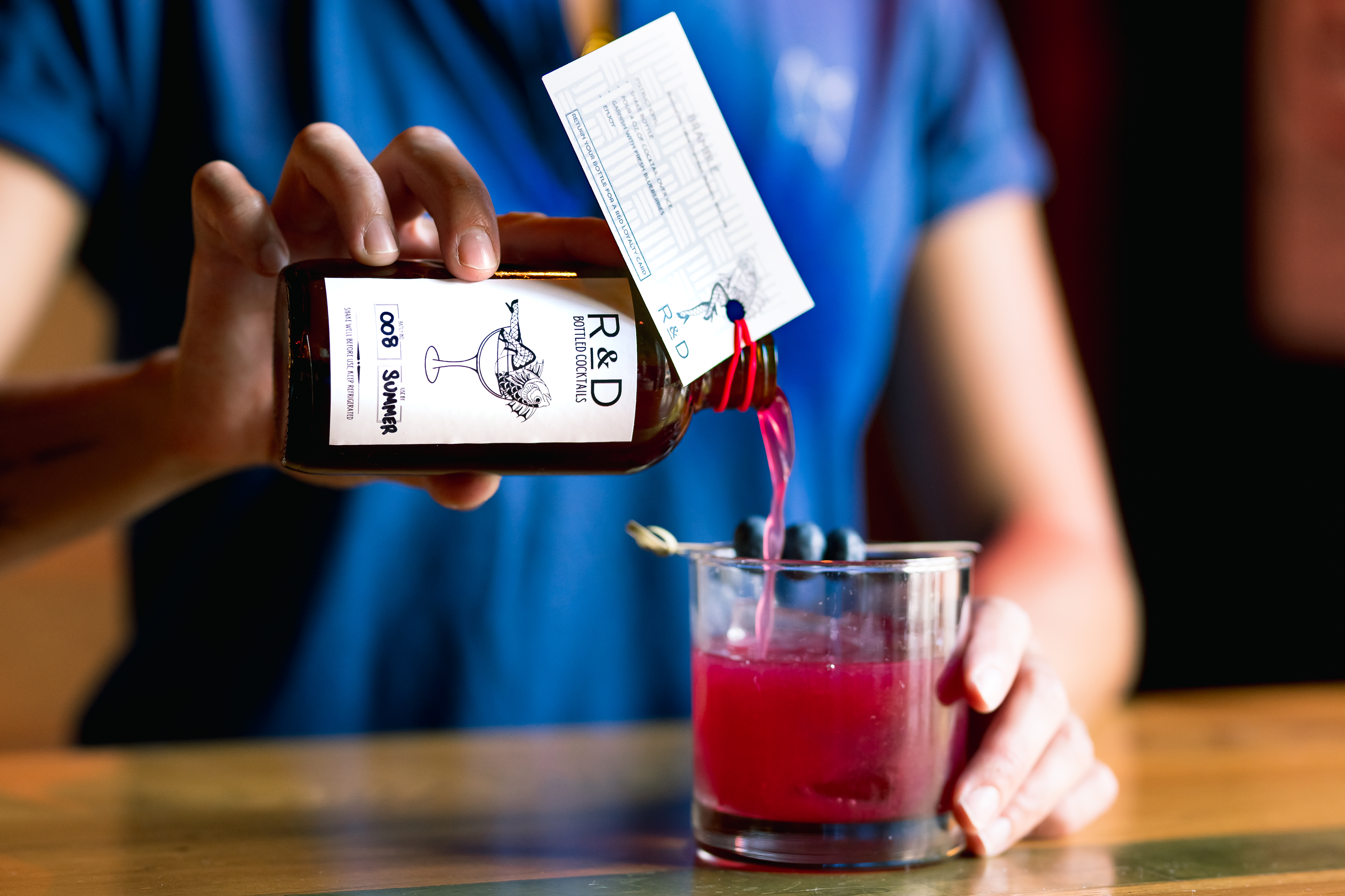 person in blue shirt pouring pink cocktail from bottle into glass