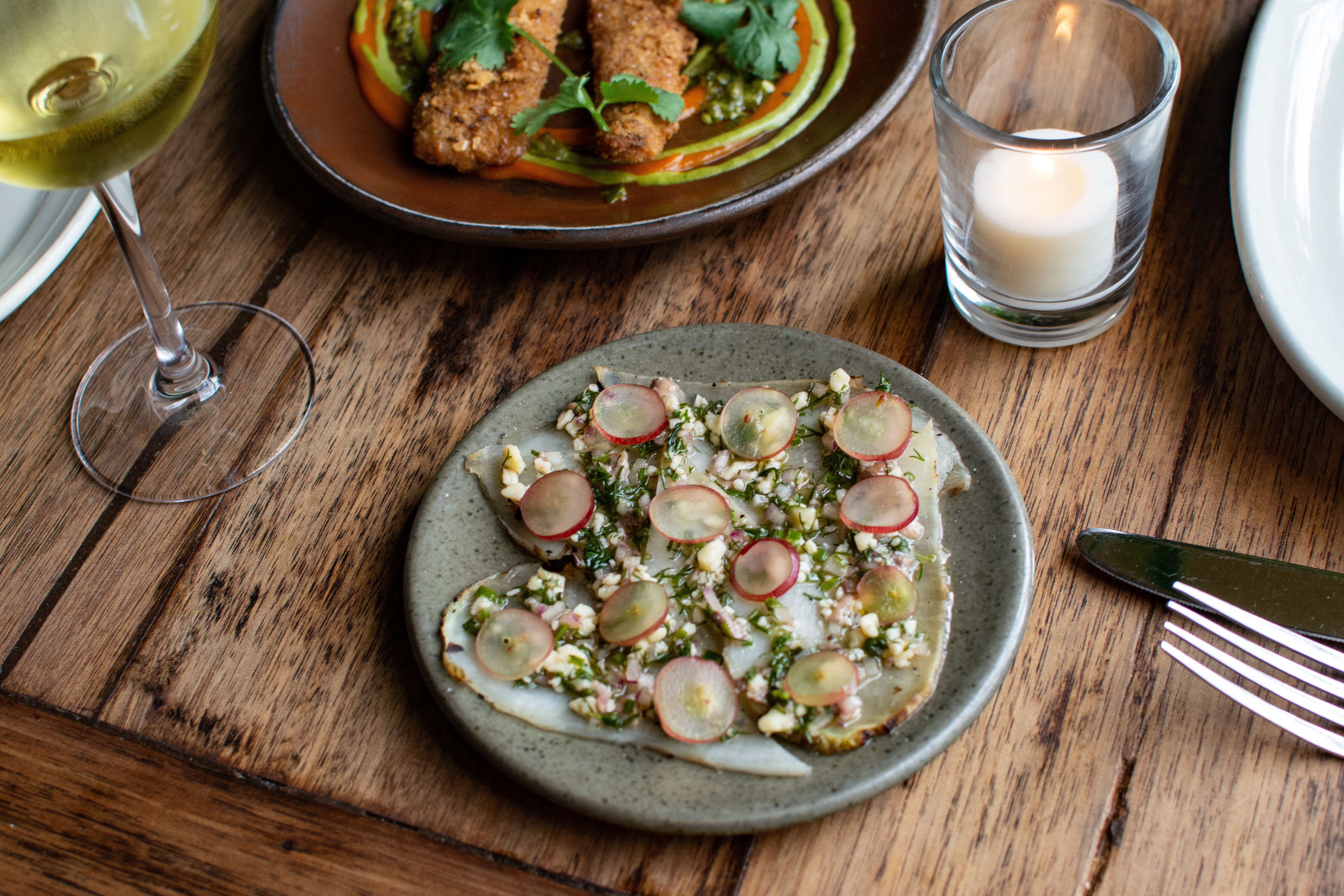 Grilled Radishes with Anchovy, Kashkaval, and Grapes, carrot schnitzel, white wine, on a wood table