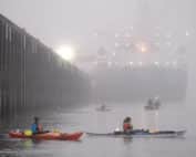 Activists in multicolored kayaks blockade a large barge--a pier is on the left side, and the sun is visible on the horizon through the gray mist.