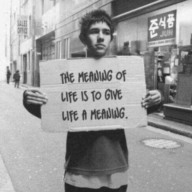Image description: A black and white photo of a person standing in the middle of a street holding a sign which says “the meaning of life is to give life a meaning”.