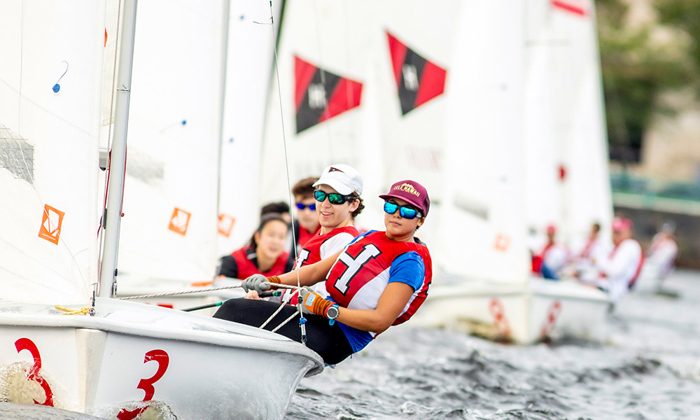 Several members of Harvard's sailing team sit together on a boat with boats lined up behind them