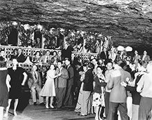 Dozens of people dressed in evening wear dance and enjoy an event inside a cave, under a rocky ceiling. Black-and-white photo.