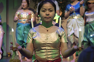 Dancers on stage in shimmering green, white, and gold costumes. A woman in front looks straight into the camera, holding a battery-powered candle in each hand.