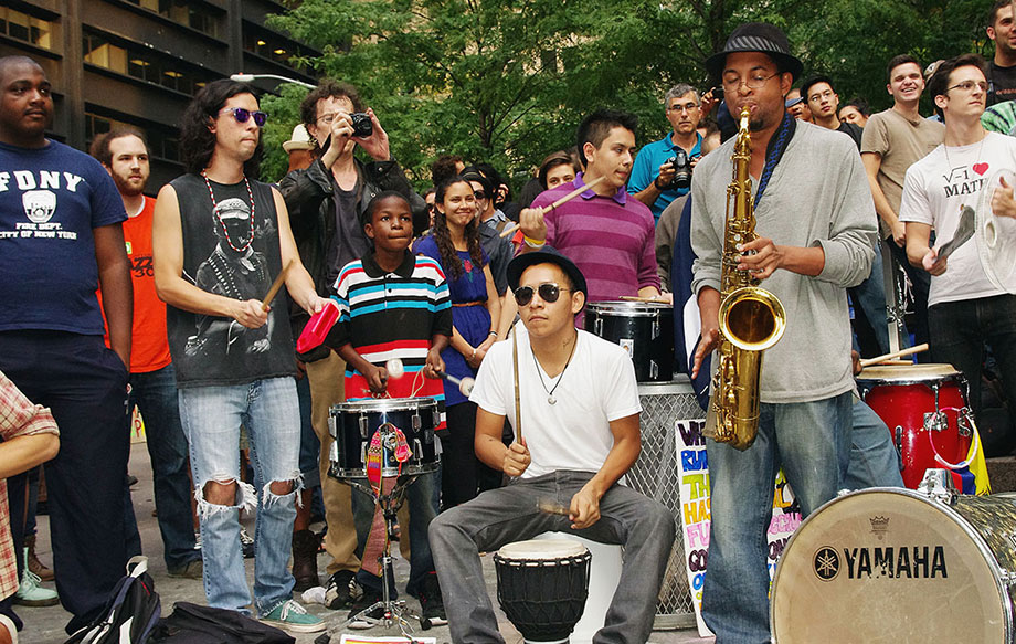 Crowd of people playing music outside, with others around them observing. Most people play some kind of percussion; one person plays saxophone.