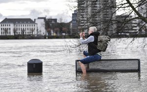 A man takes a picture with his smartphone while sitting on a bench at a the flooded bank of the Rhine river in Cologne, Germany, Wednesday, Feb. 3, 2021.