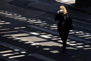 A woman wearing a mask walks beneath elevated subway tracks that cast a pattern of light on a Brooklyn street during the coronavirus pandemic, Monday, April 6, 2020 in New York