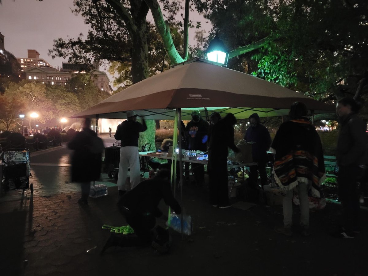 A canopy up in washington square park. People underneath it distributing water and pizza and other food