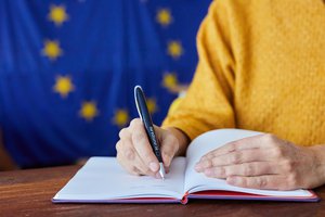 A person writing in a notebook with an European Union pen and the EU flag in the background