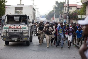 Police officers and protesters take cover behind a police truck as shots ring out during a protest to demand the resignation of Haiti's president Jovenel Moise on the 216th anniversary of Battle of Vertieres in Port-au-Prince, Haiti, Monday, Nov. 18, 2019.