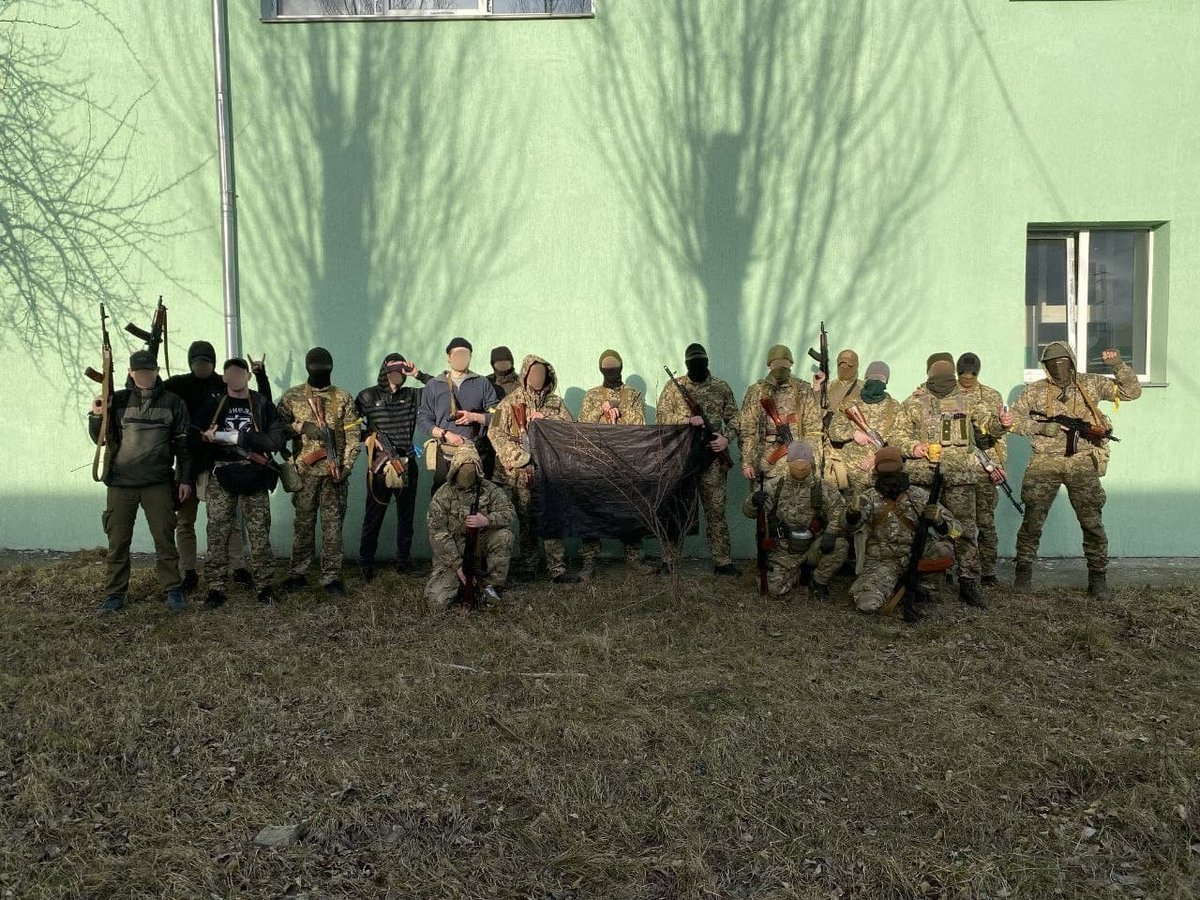 A photograph of nineteen people, some wearing camouflage, some not, holding assault rifles and posing with a black flag representing anarchism, the struggle for a world without domination.