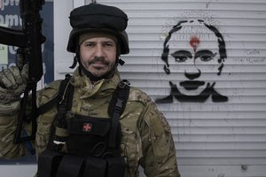 A Ukrainian serviceman poses next to a graffiti depicting Russian President Vladimir Putin and the words 'Glory to Ukraine" on the blinds of a battle damaged shop in Stoyanka, Ukraine, Sunday, March 27, 2022.