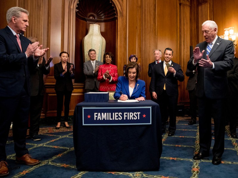 Nancy Pelosi, wearing a blue suit, sits at a table signing a bill. A sign reading "families first" is on the table. A group of officials surrounding her applauds.