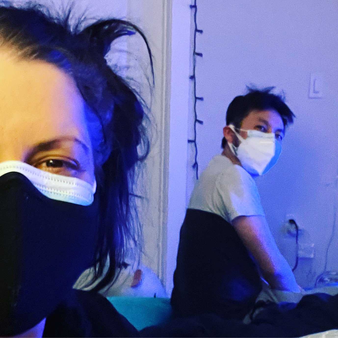 Sara, a white Disabled brunette, is taking a selfie with her face in two masks in the foreground. In the background is her partner, Kal, a Hmong American man wearing 2 kn95 masks.