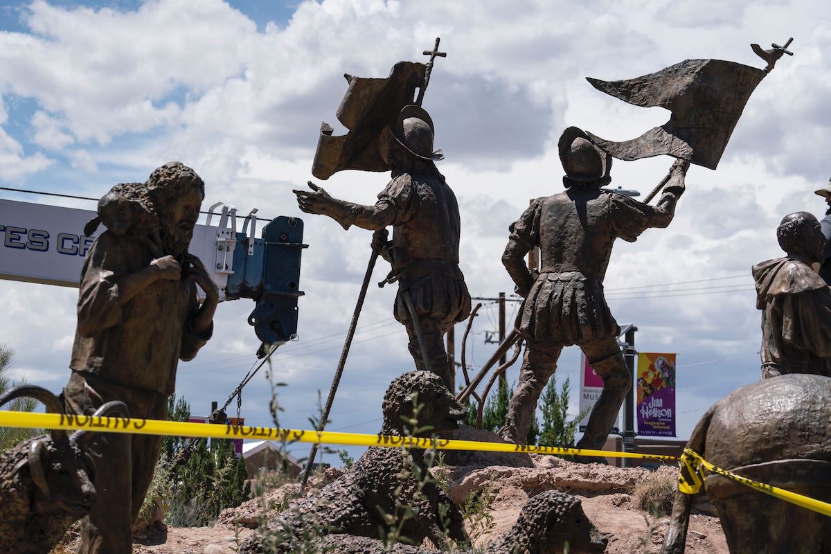 Statues of Spanish conquistadors are surrounded by a perimeter of caution tape. The arm of a heavy crane stands in the background.