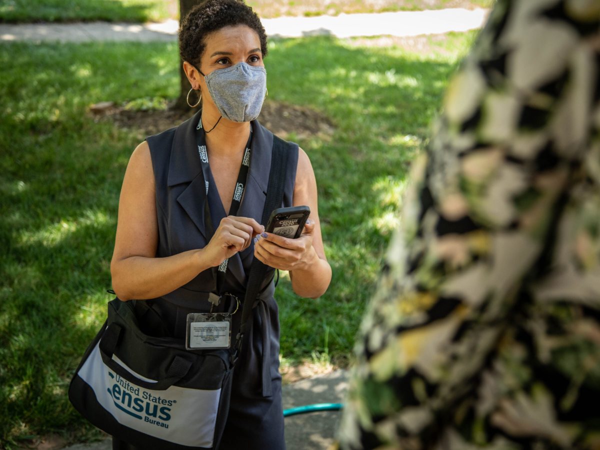 A census worker wearing a facemask and carrying a phone and branded bag talks to a person in the foreground.